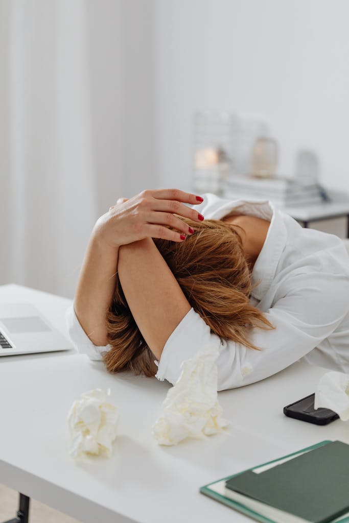 A woman in distress sits at a desk with head in hands, surrounded by tissue papers.