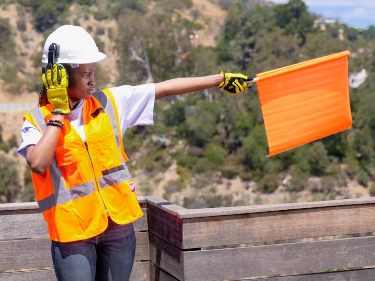 Woman in safety gear signaling with an orange flag outdoors.
