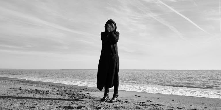 Black and white image of a person in long coat expressing sadness on a beach.