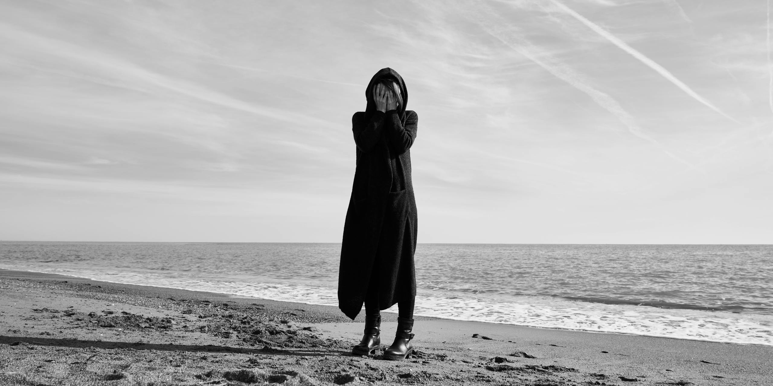 Black and white image of a person in long coat expressing sadness on a beach.